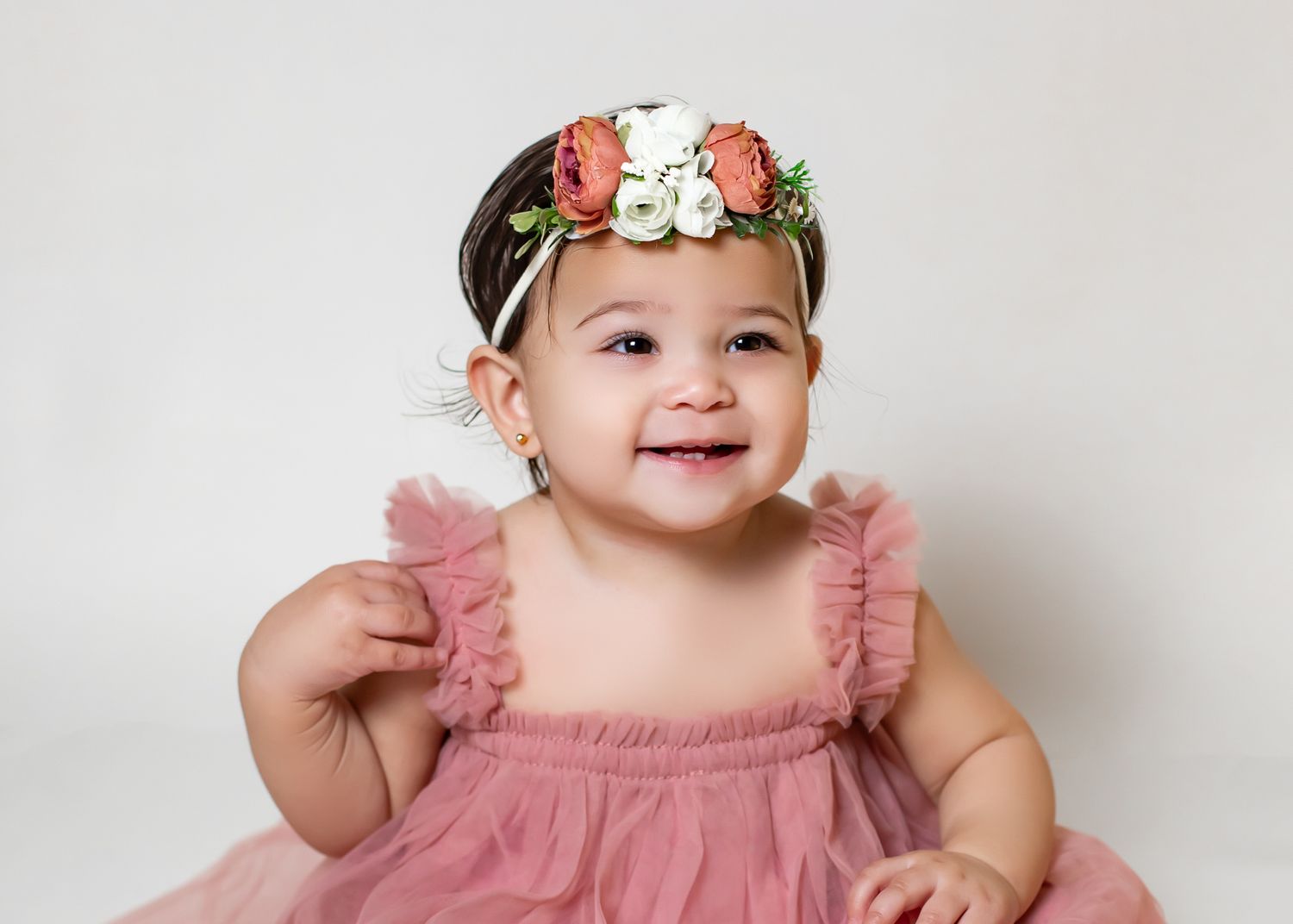Happy baby girl in pink ruffle dress and floral crown smiling brightly on white background.