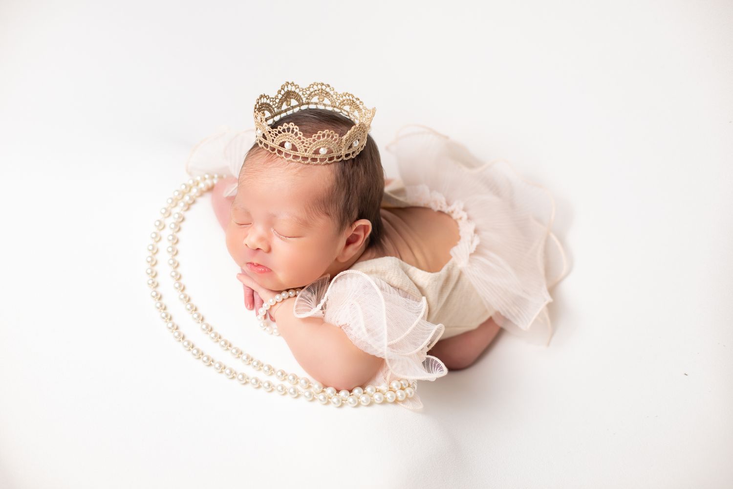 Newborn baby girl sleeping in white outfit with gold crown and pearl necklace on white background.
