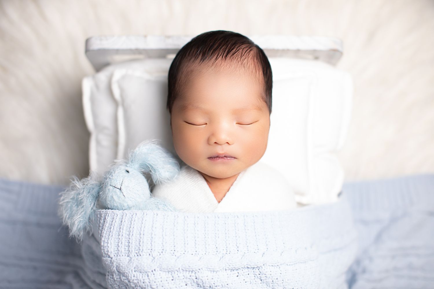 Sleeping newborn baby in white swaddle on blue knit blanket with a small blue stuffed bunny toy.