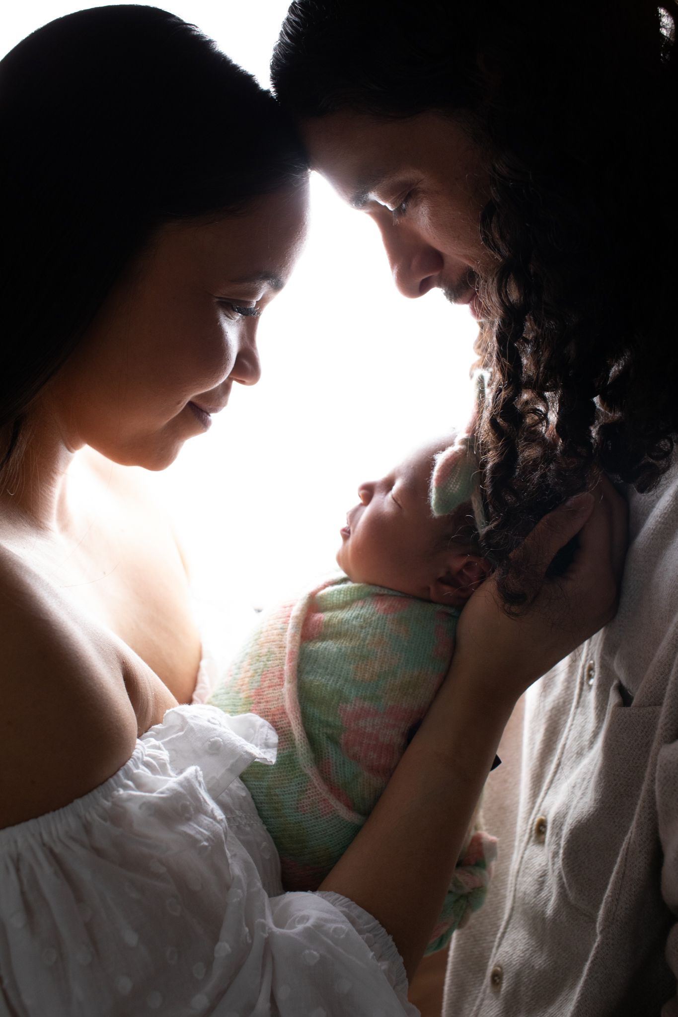 Silhouetted parents gazing lovingly at their newborn baby held between them in soft backlight.