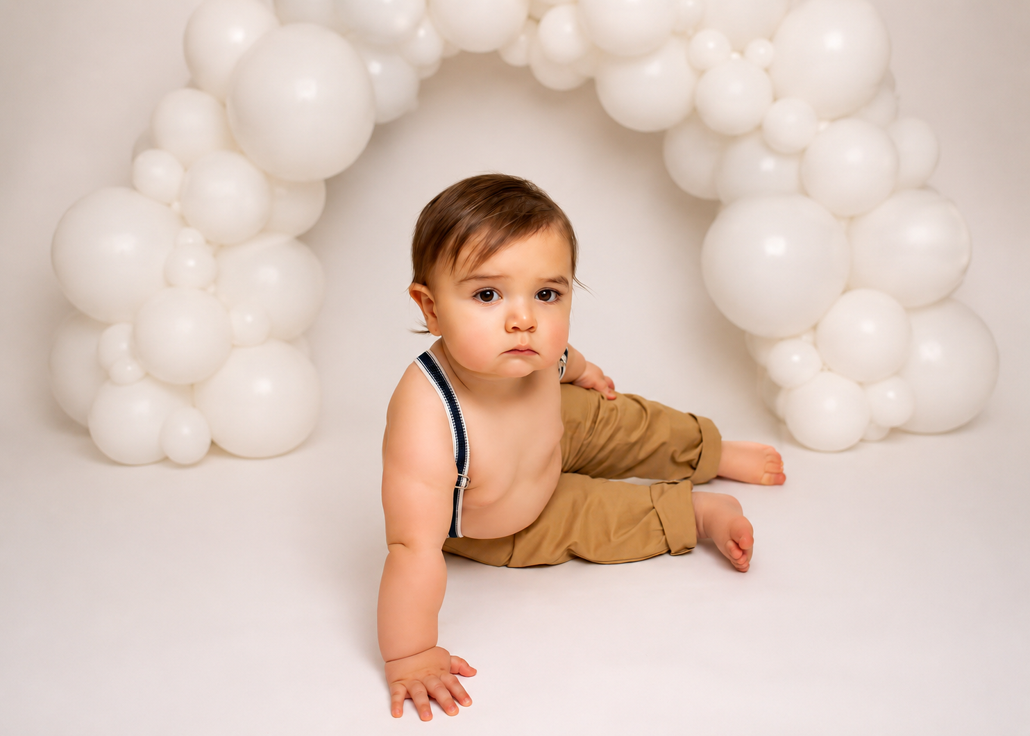 Baby boy in tan overalls sits before a white balloon arch on a white backdrop in a professional photo.