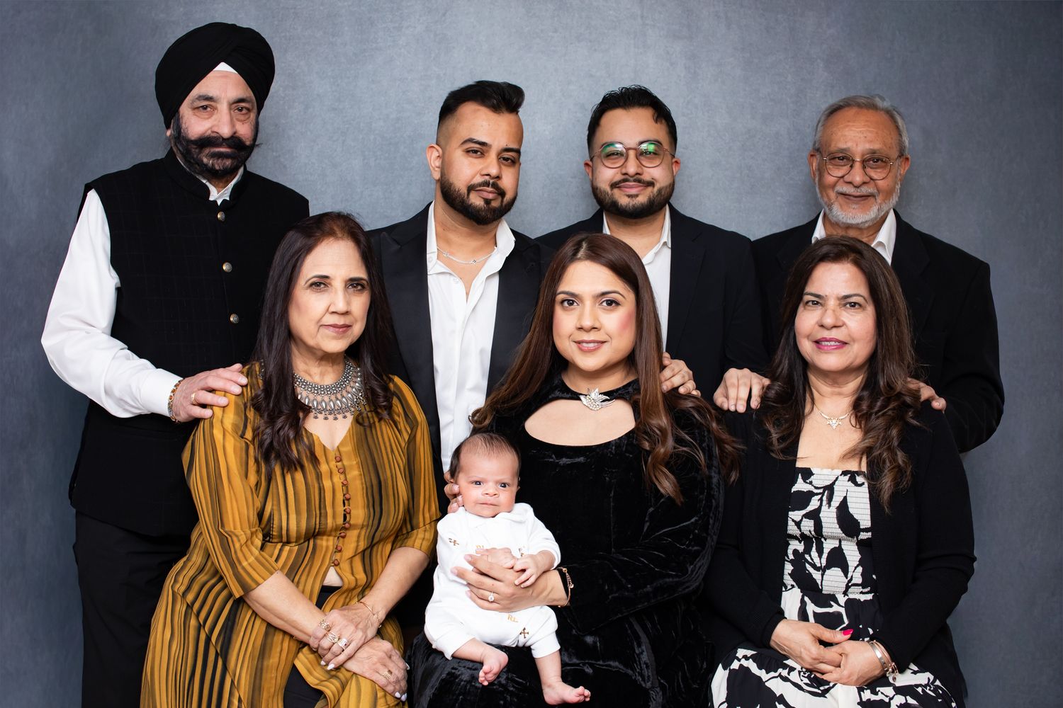 Multi-generational South Asian family portrait with adults and a baby, dressed in formal attire, posing together indoors.