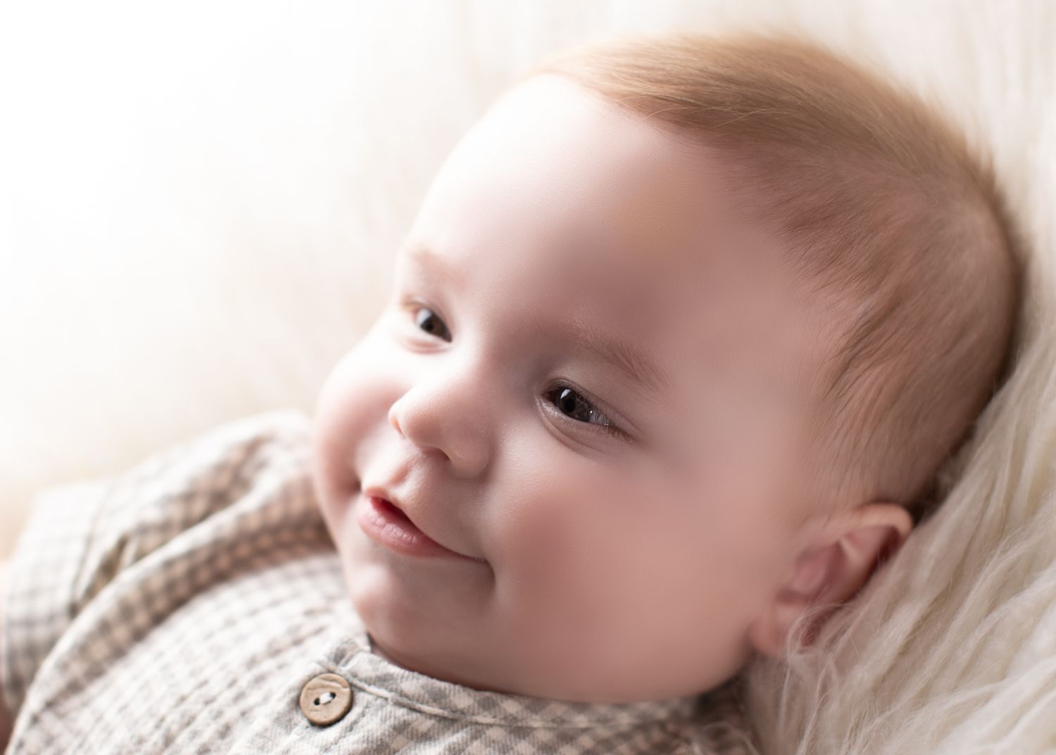 Adorable smiling baby in a brown plaid outfit lying on a soft white fluffy surface.
