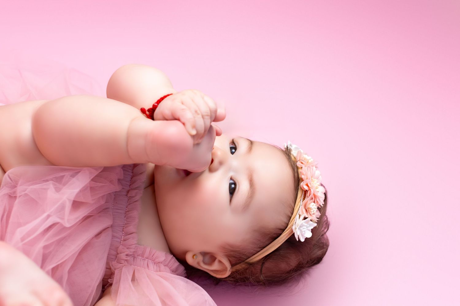 Adorable baby girl in pink tutu dress and floral headband lying on pink background, looking up curiously.