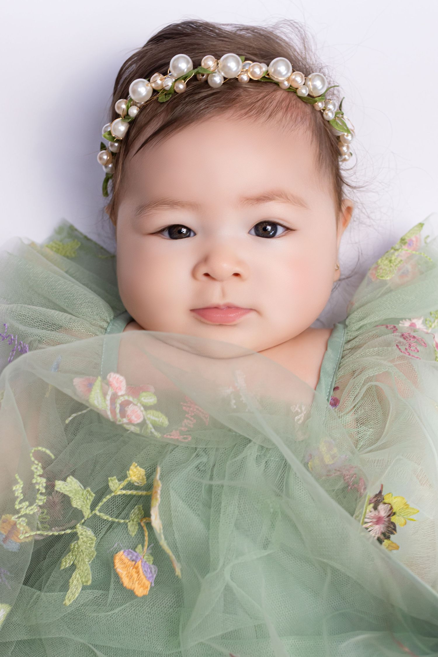 Adorable baby girl wearing a pearl headband and green floral tulle dress, lying down with a sweet expression.