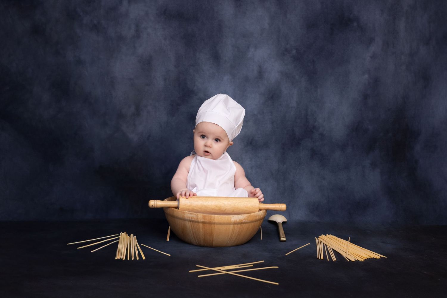 Baby in chef hat sits in wooden bowl with rolling pin and pasta on dark background.