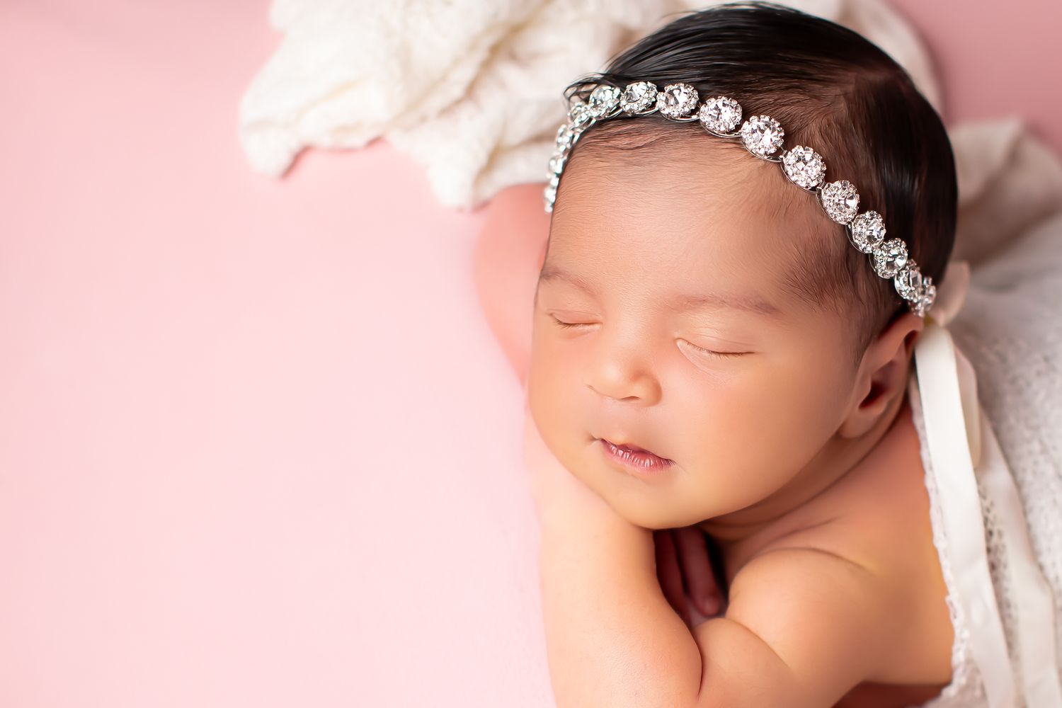 Peaceful newborn baby sleeping on pink background wearing elegant rhinestone headband.