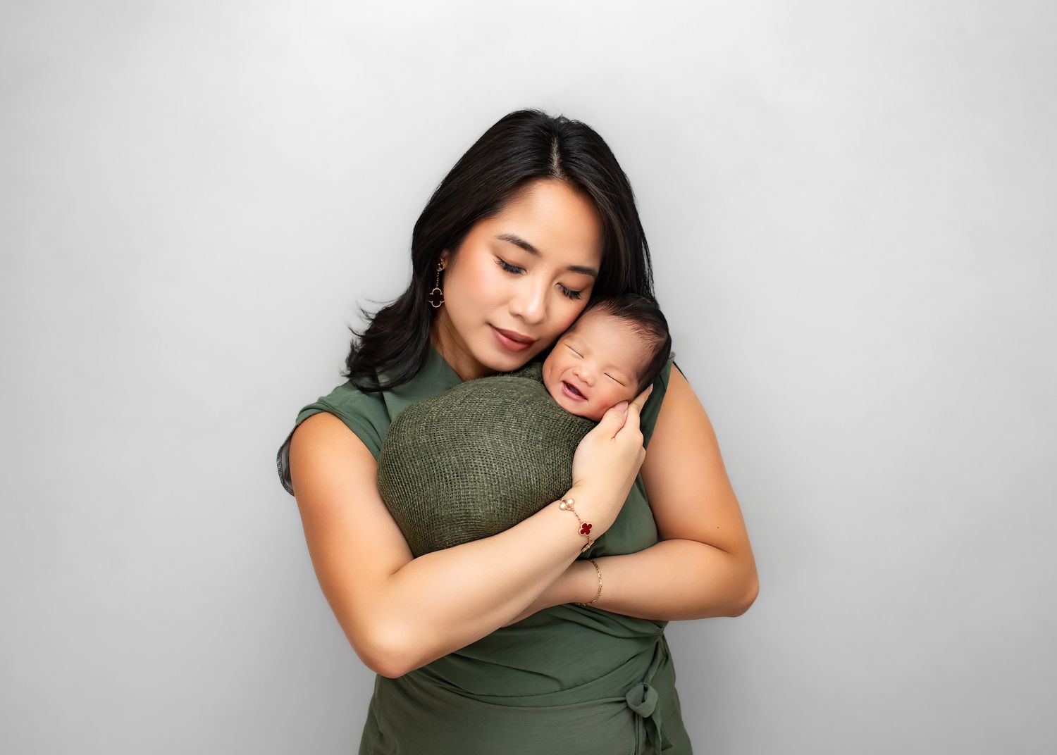 Mother in green dress cradling sleeping newborn baby against gray studio background.