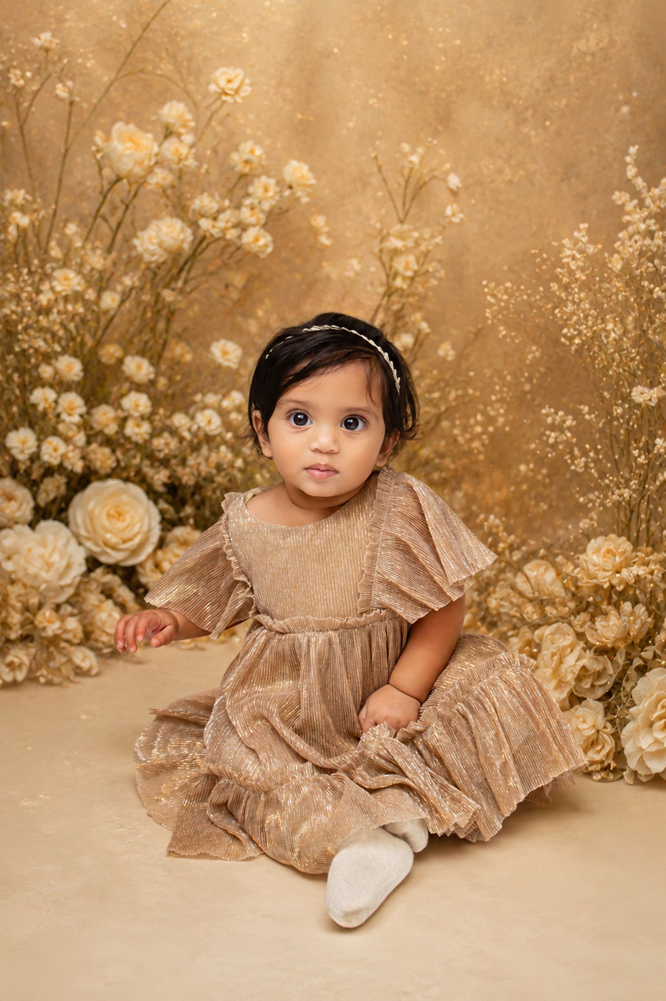 Cute baby girl in a golden ruffle dress sitting among white flowers and roses on a warm golden backdrop.