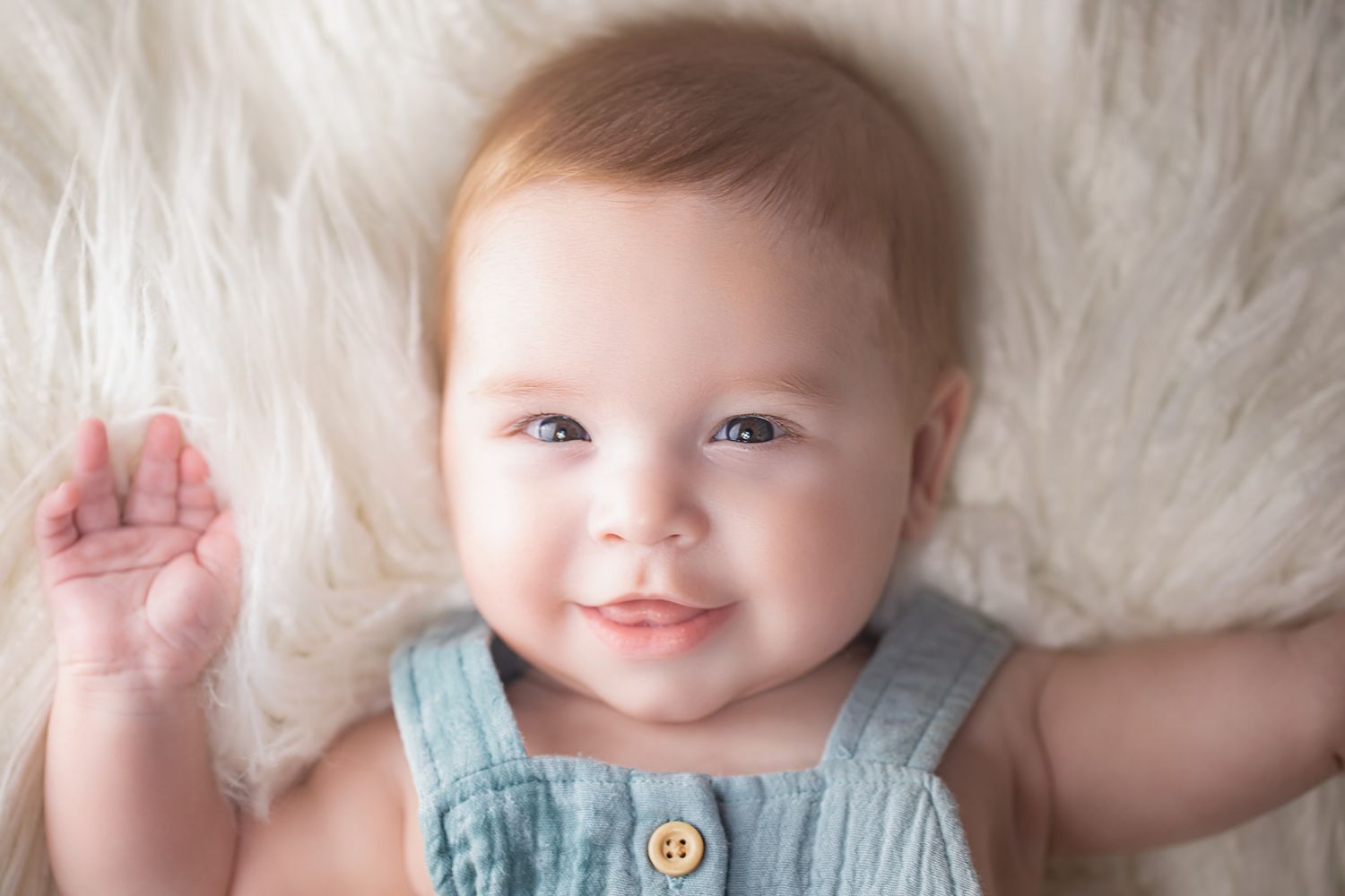 Smiling baby in a teal sleeveless romper lying on a fluffy white fur surface.