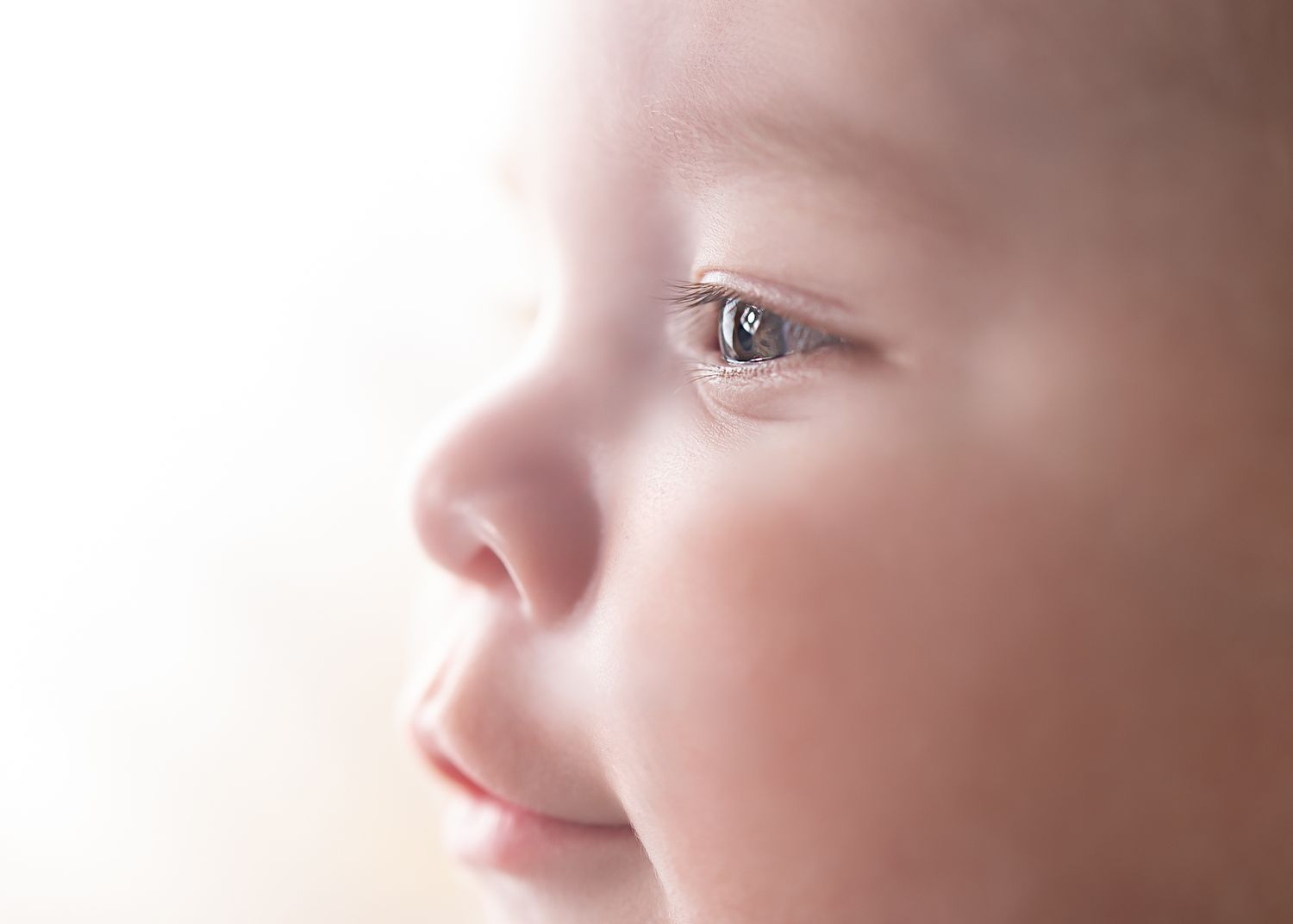 Close-up side profile of a baby with bright eyes gazing upward on a light background.