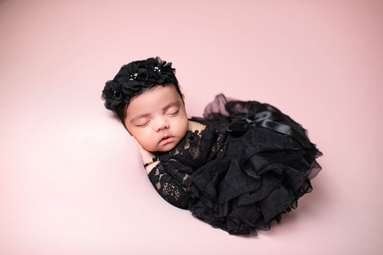 Sleeping newborn baby girl in black lace dress and floral headband on pink background.