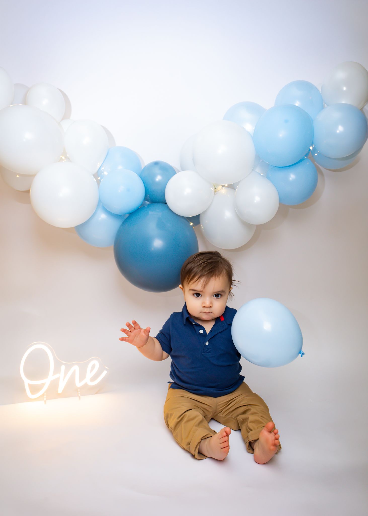 Baby boy first birthday photo with blue balloon garland and glowing One neon sign backdrop.