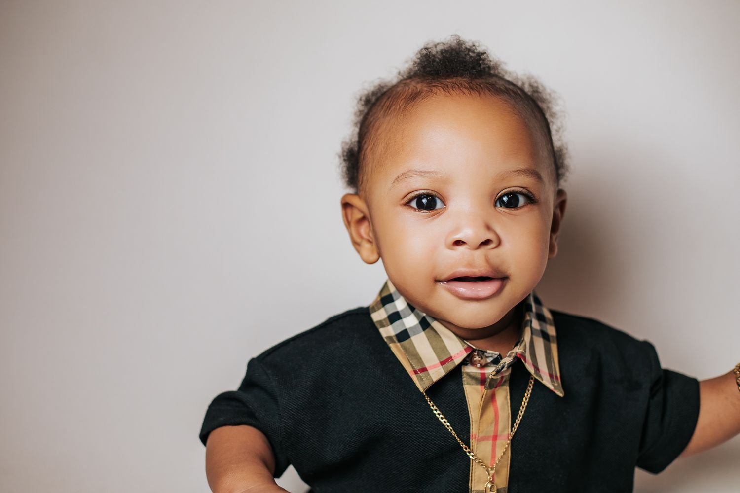 Charming baby boy in a black Burberry collared shirt smiling against a light gray background.