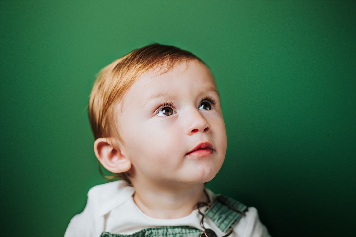 Adorable toddler boy with red hair looking up curiously against a vibrant green background.