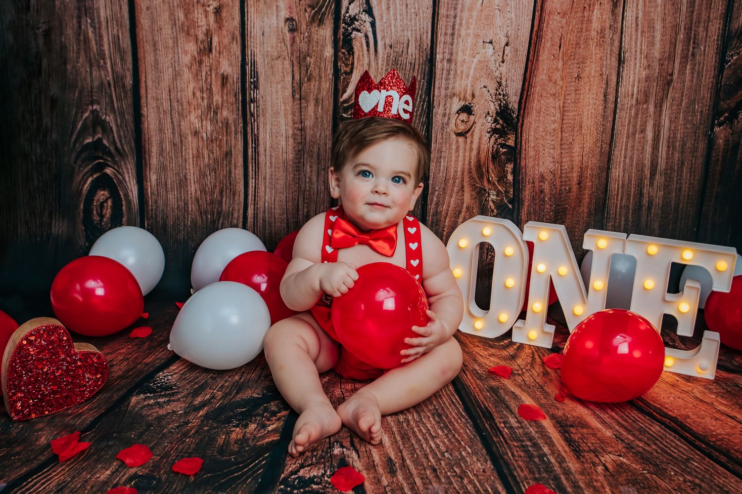 Baby in red polka dot outfit with 'One' crown beside balloons and marquee letters on wood backdrop.