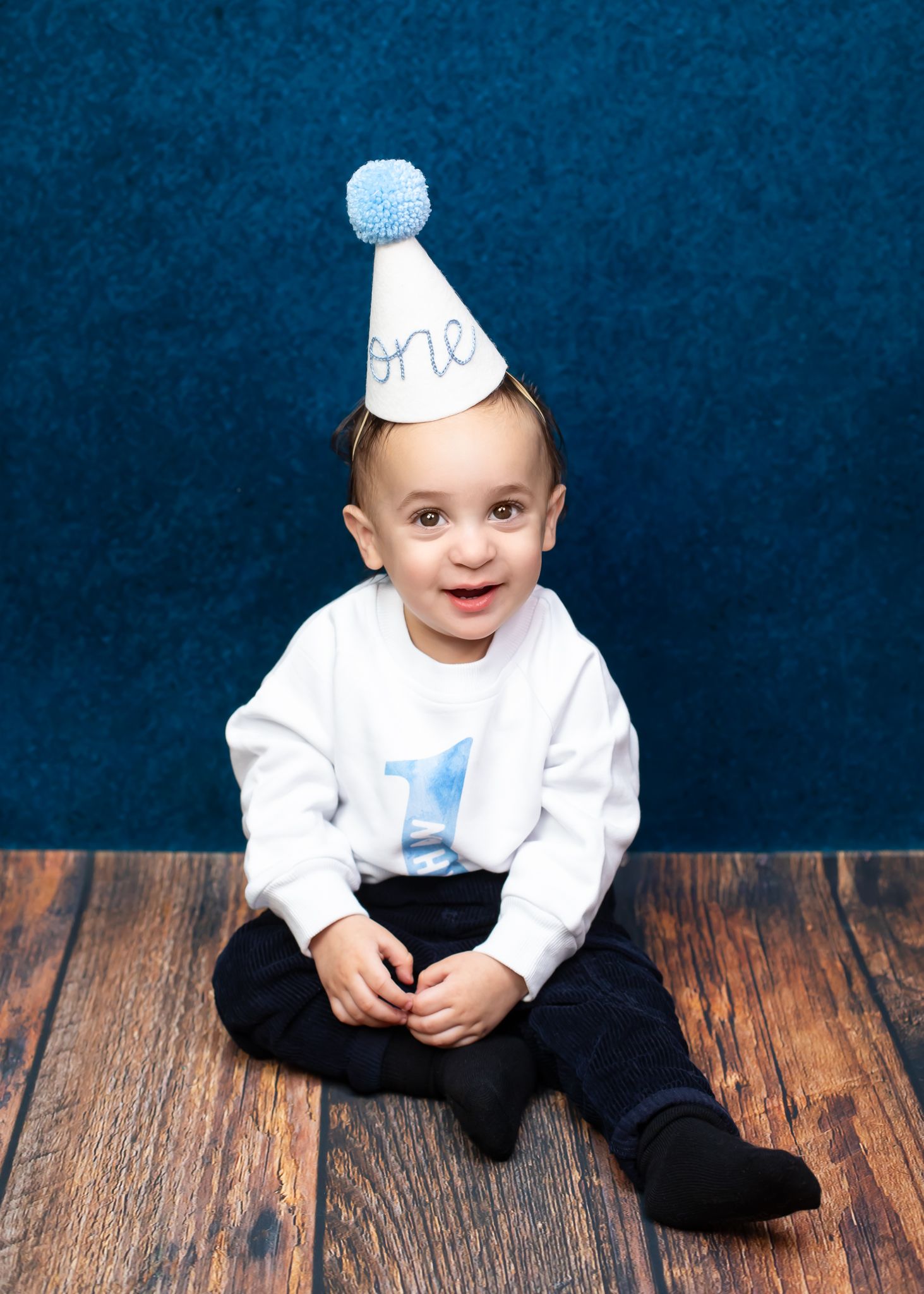 Smiling baby boy in white shirt and blue party hat that reads one sitting on wood floor for first birthday.