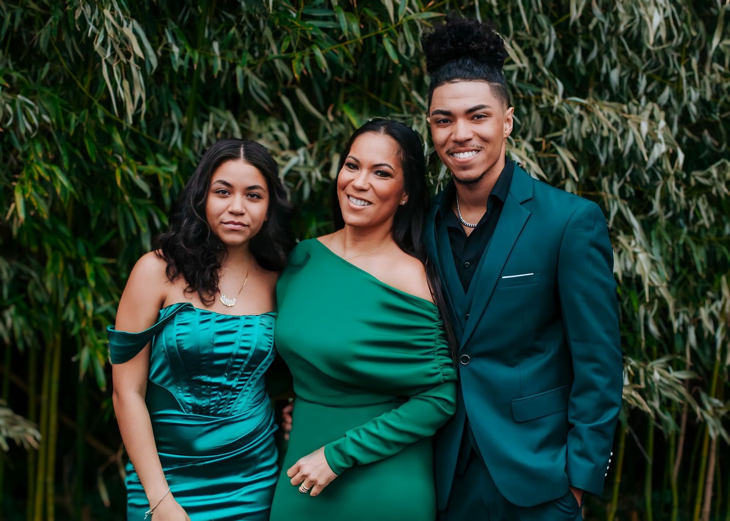 Three people in coordinating green outfits posing together in front of lush bamboo greenery.
