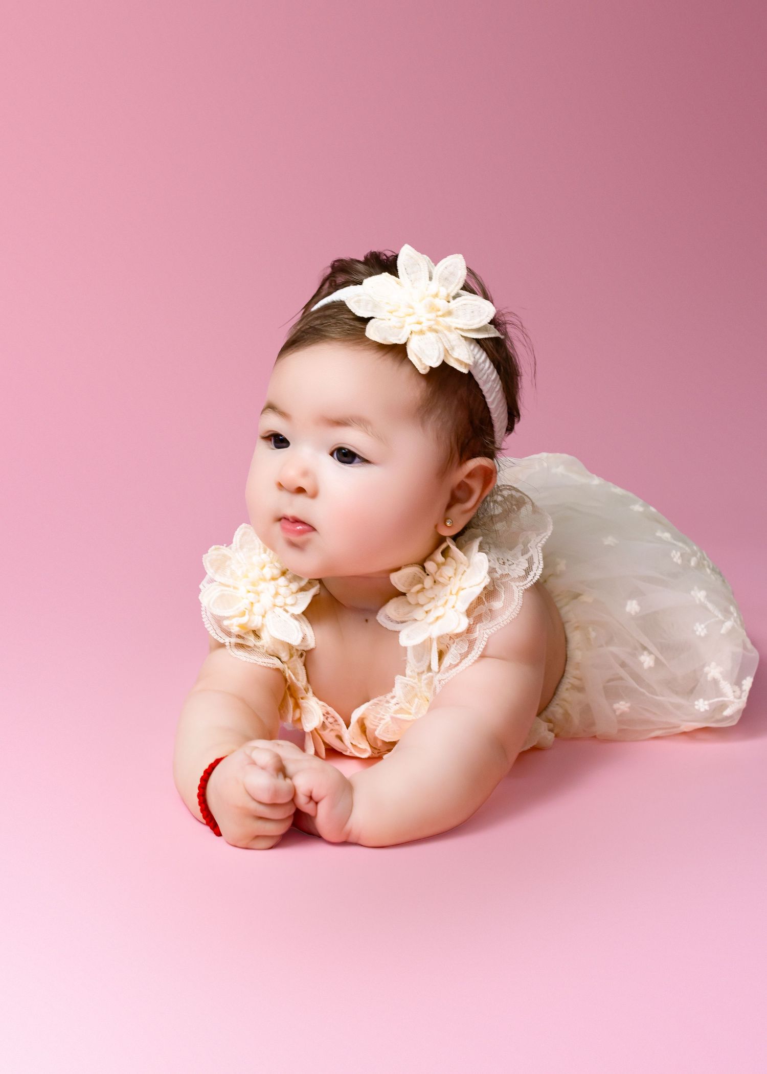 Adorable baby girl in white lace dress and floral headband poses on pink background.