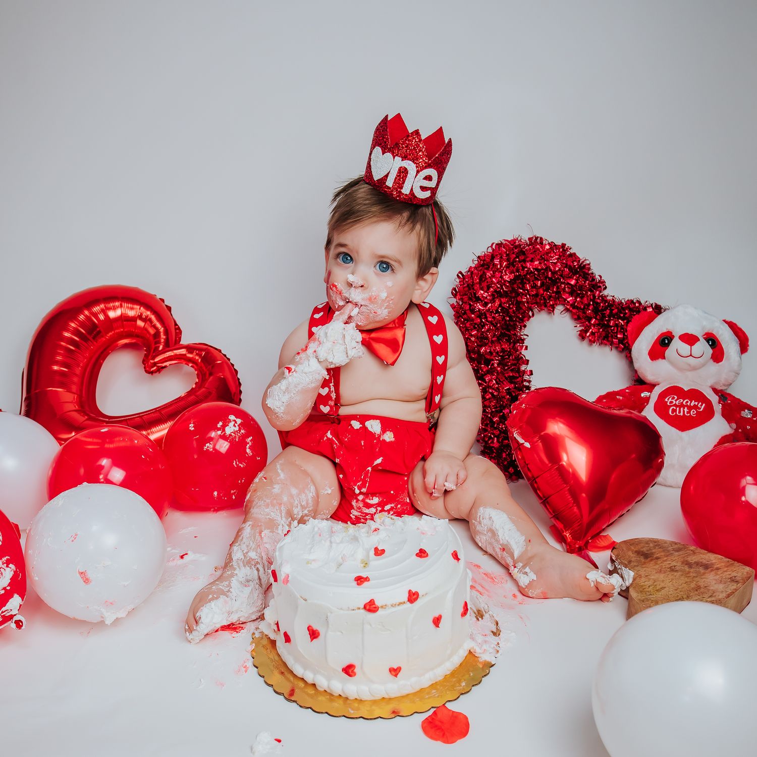 One-year-old baby in red outfit enjoying Valentine's Day cake smash with heart balloons and teddy bear.