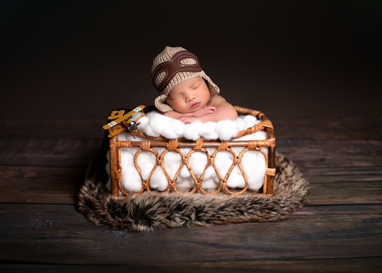 Sleeping newborn baby in brown knit hat resting in a wicker basket with fur trim.