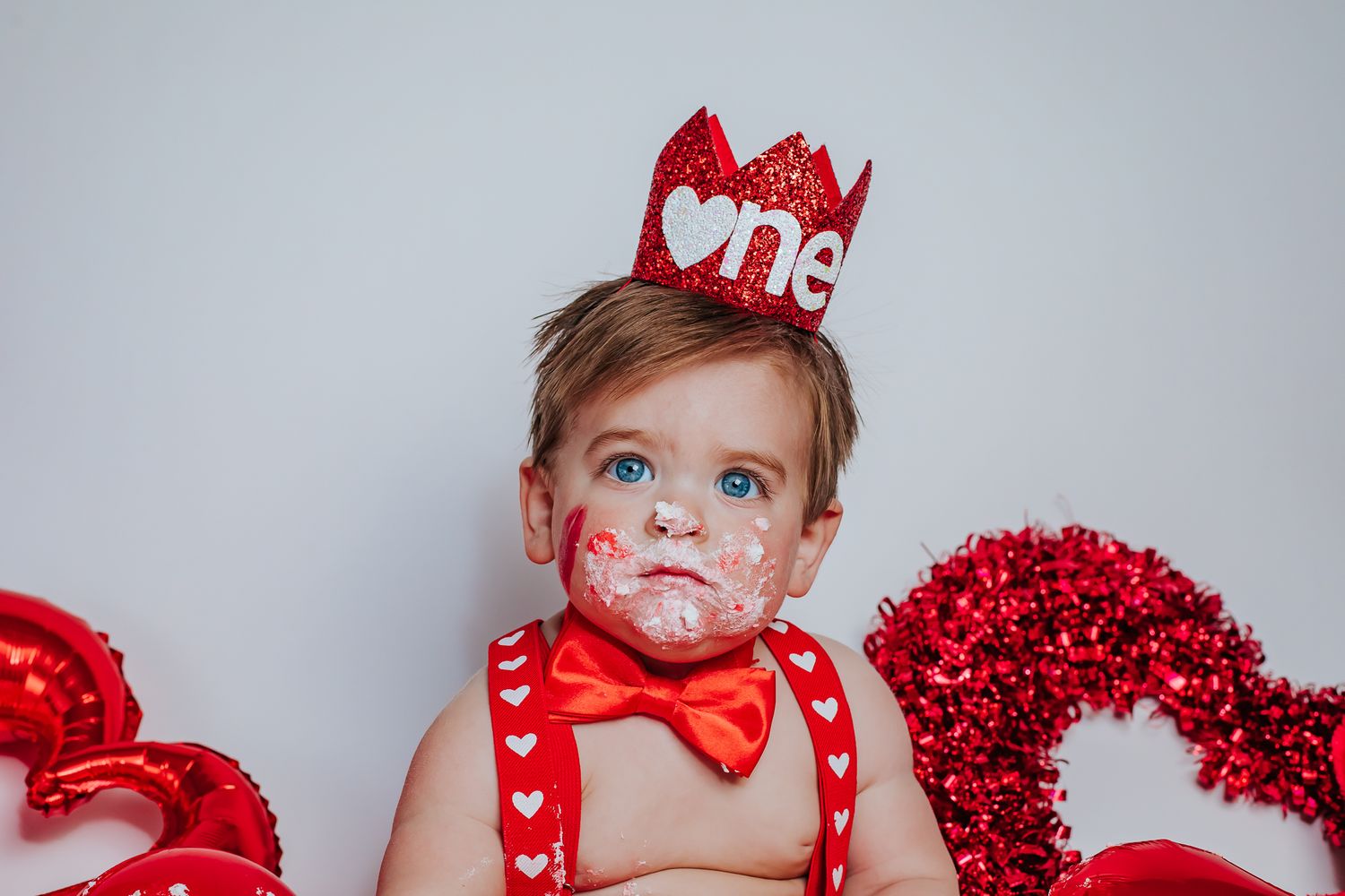 Close-up of baby with cake-covered face wearing glittery red 'One' crown for Valentine's Day birthday.