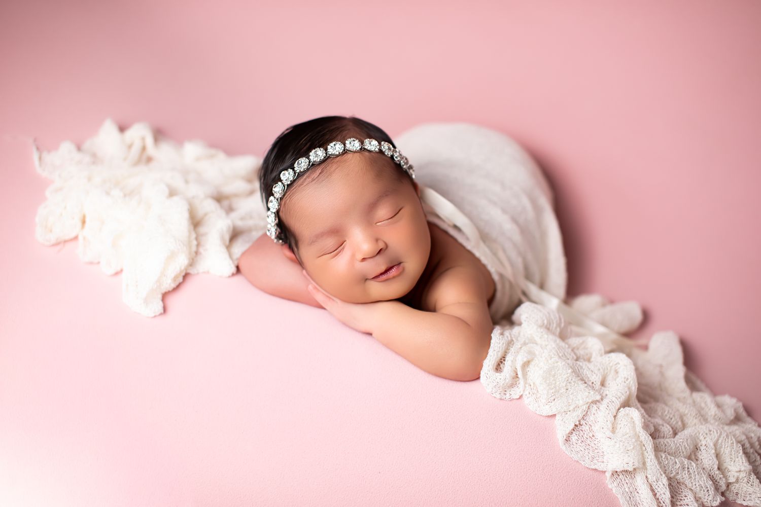 Smiling newborn baby girl with pearl headband wrapped in white lace on pink background.