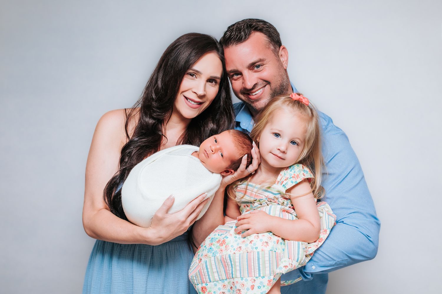 Smiling family posing together with a swaddled newborn baby and young toddler girl in a studio.