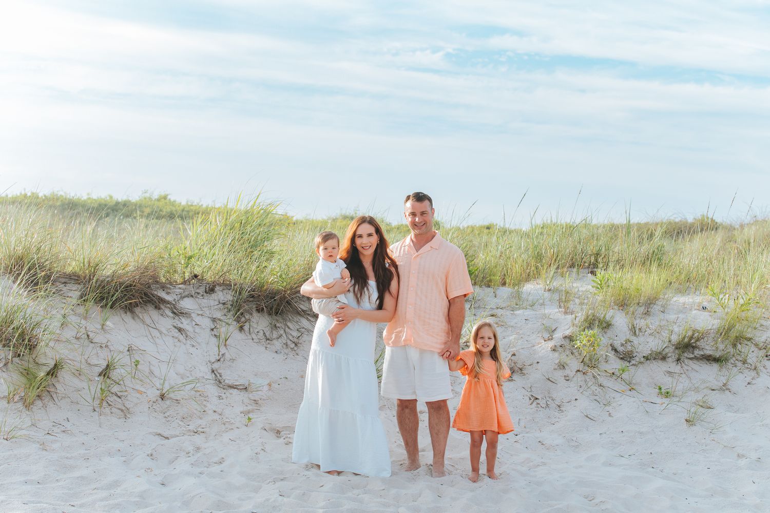 Happy family of four standing together on sandy beach near grass dunes in coordinating outfits.