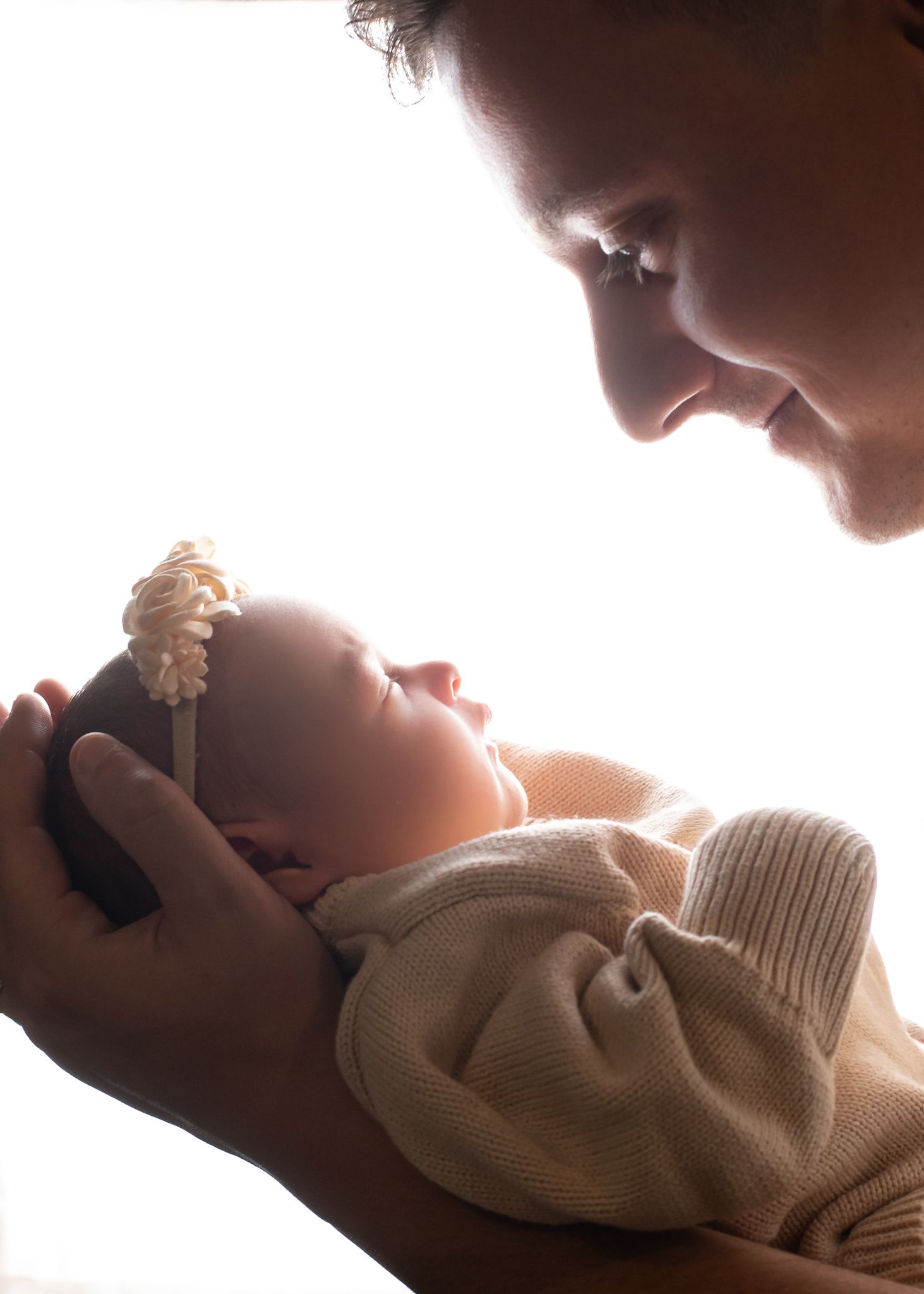 Silhouette of parent gazing lovingly at sleeping newborn baby held in hands against bright light.