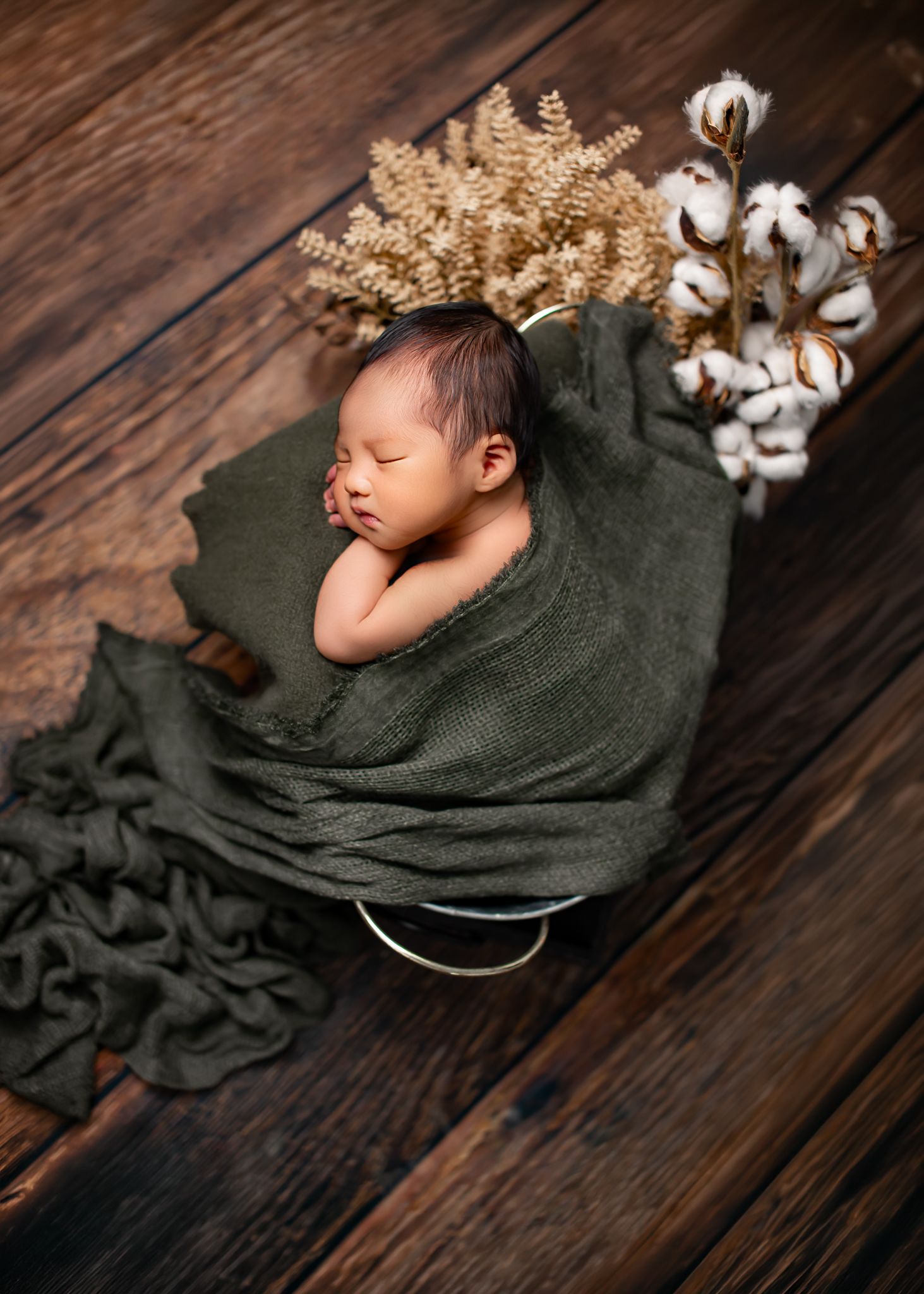 Sleeping newborn baby wrapped in olive green cloth inside a bucket with dried flowers on wood floor.