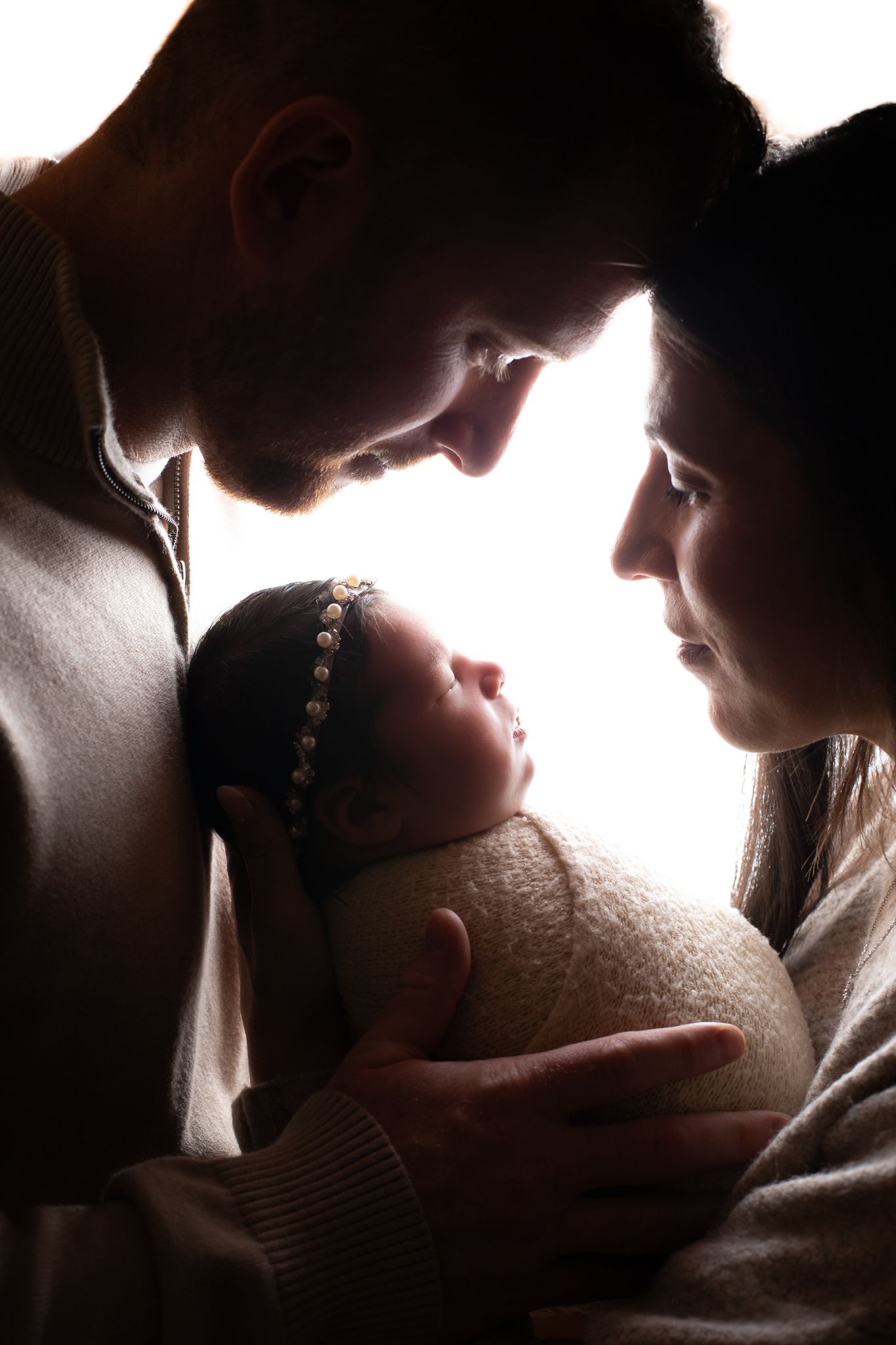 Silhouette of parents gazing at their swaddled newborn baby in soft backlight.