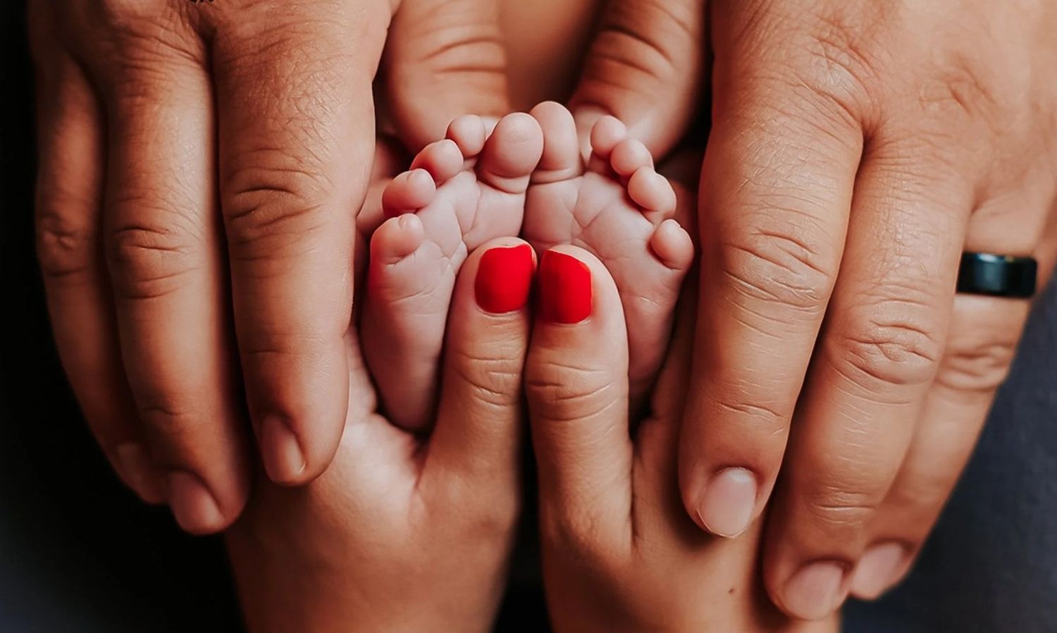Two adult hands cradling tiny newborn baby feet with red painted toenails in a tender family moment.