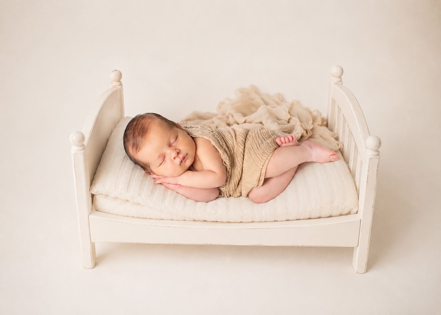 Smiling newborn baby in beige wrap sleeping peacefully on a small white miniature bed prop.