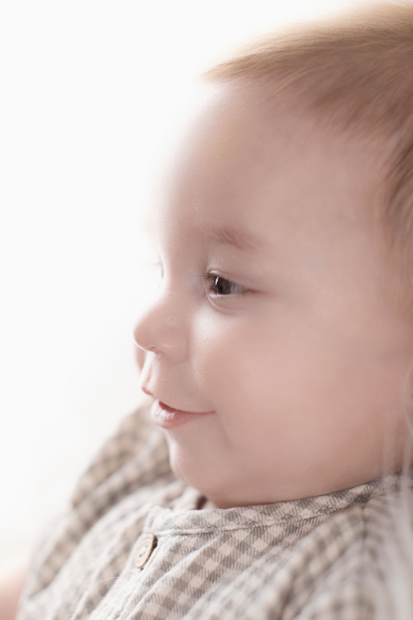 Side profile of a smiling baby in a plaid outfit against a bright white background.