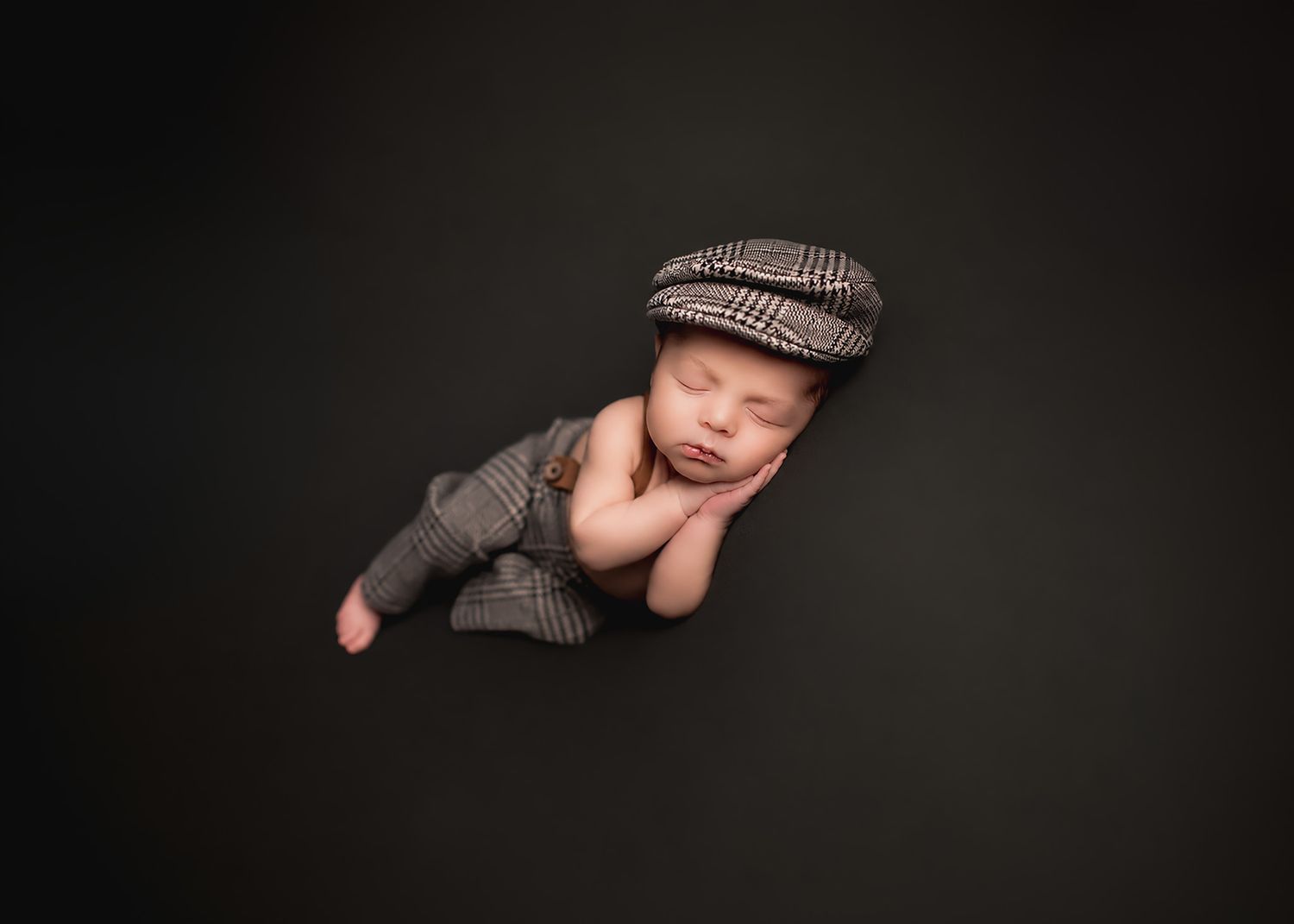 Sleeping newborn baby wearing a gray plaid flat cap and matching outfit, posed on a dark background.