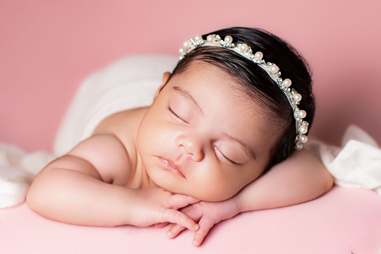 Sleeping newborn baby girl with dark hair wearing pearl headband on soft pink backdrop.