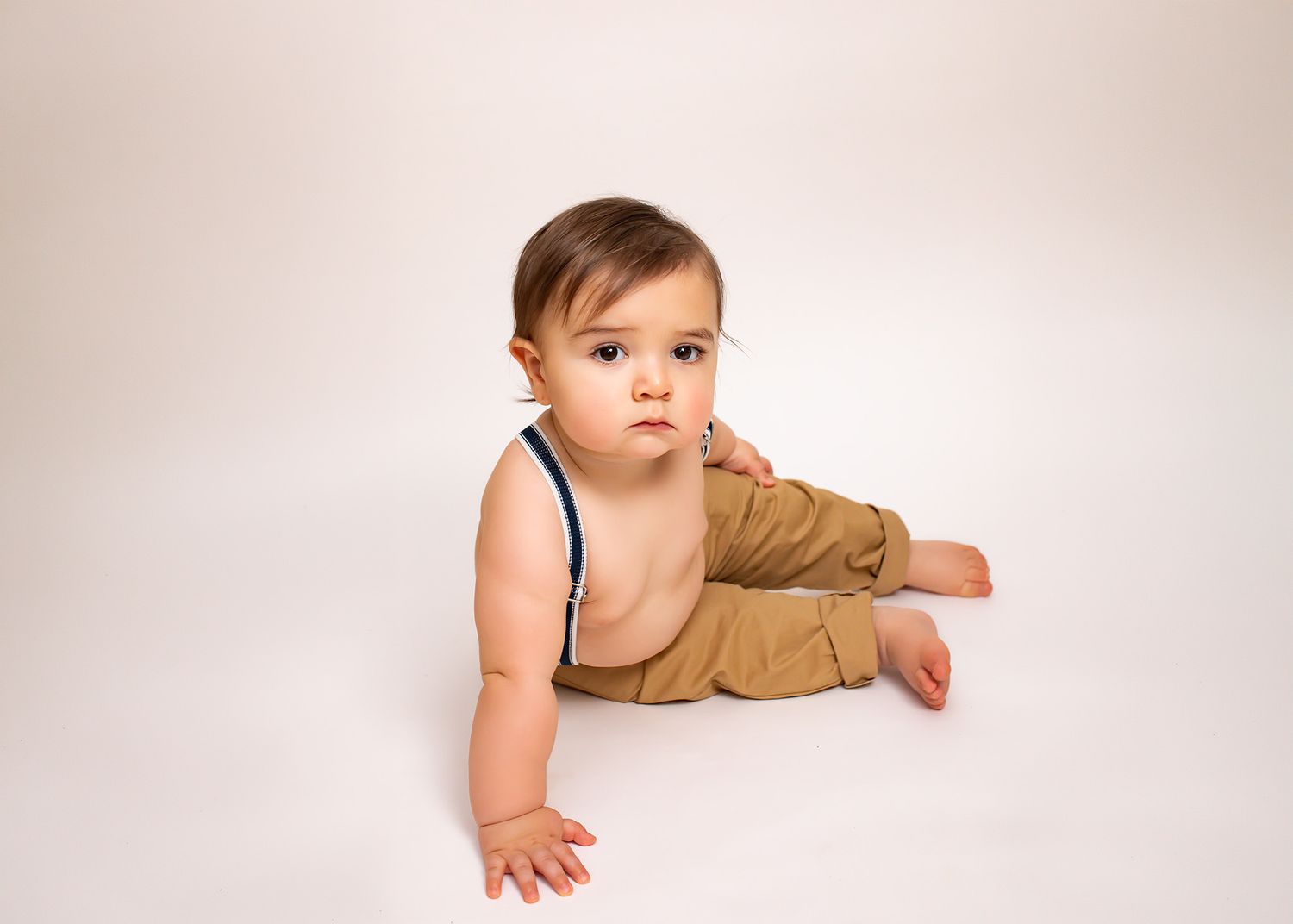 Cute baby boy in tan suspender pants sitting on white floor during professional photo session.