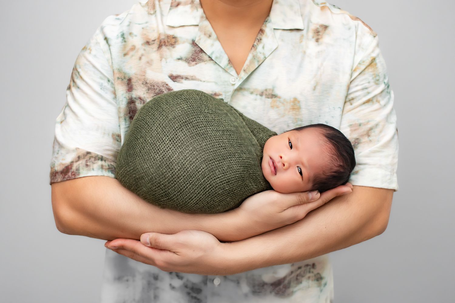 Parent cradling a swaddled newborn baby in a green wrap against gray background.