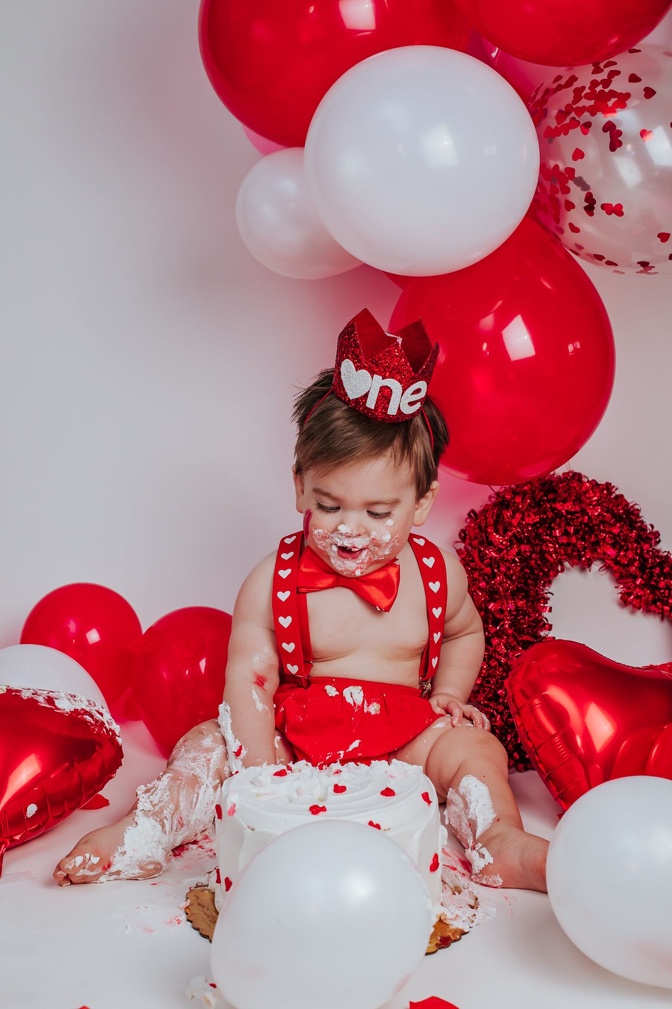 Baby wearing red one crown smashing white heart cake with red and white Valentine's Day balloon backdrop.