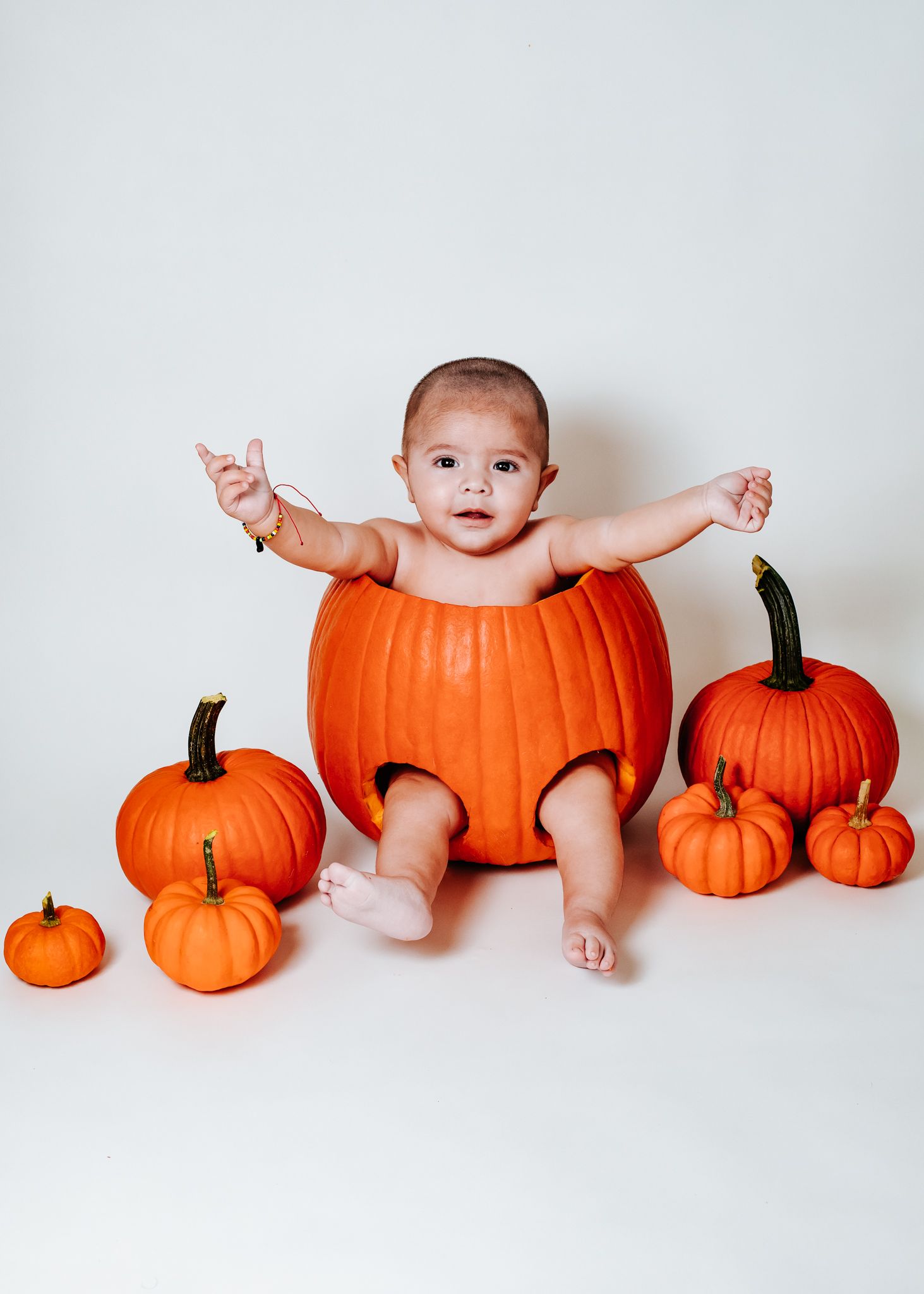 Smiling baby sitting inside large orange pumpkin surrounded by small pumpkins on white background.
