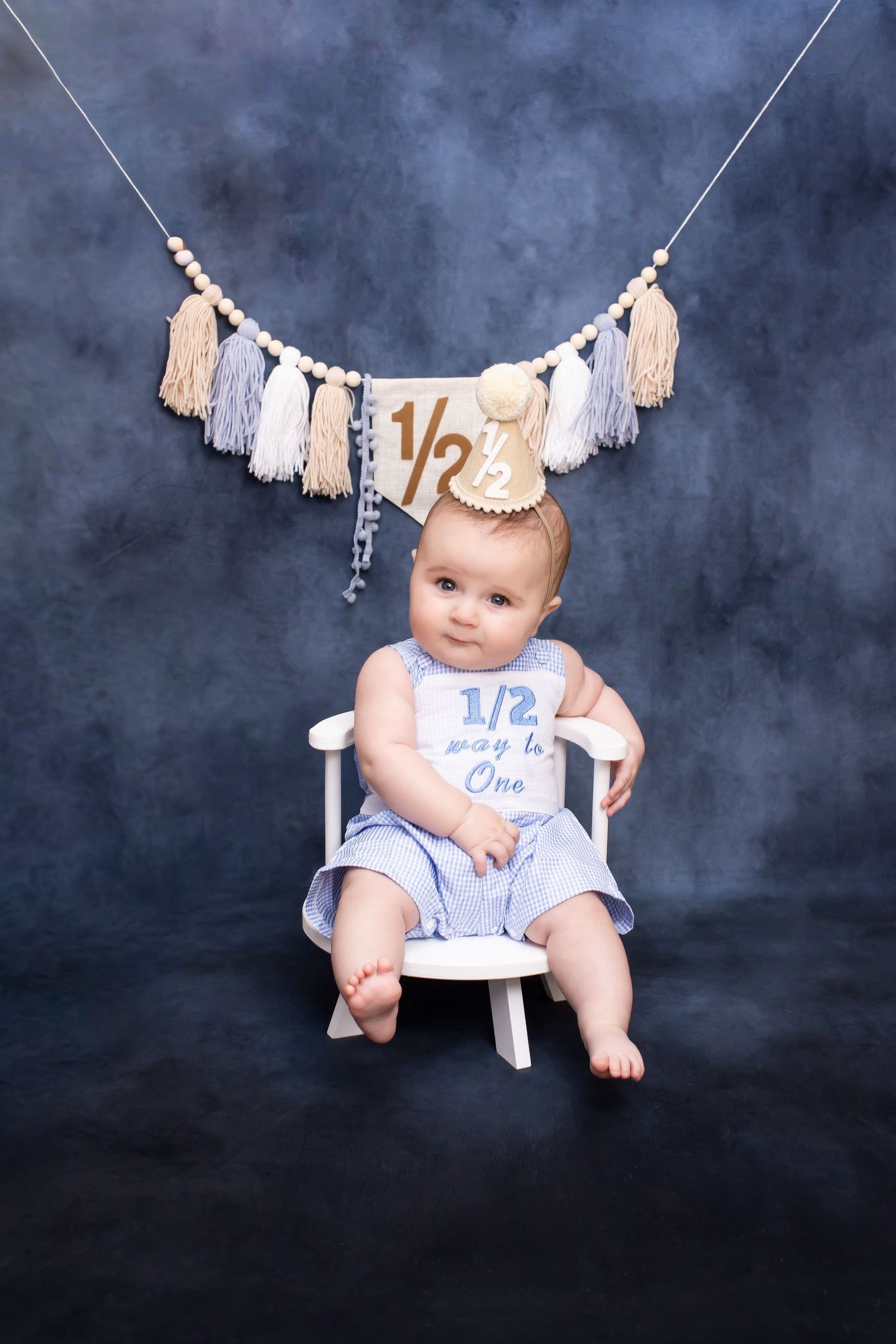 Baby in blue romper sits in white chair wearing party hat, celebrating half birthday with decorative banner and tassels.