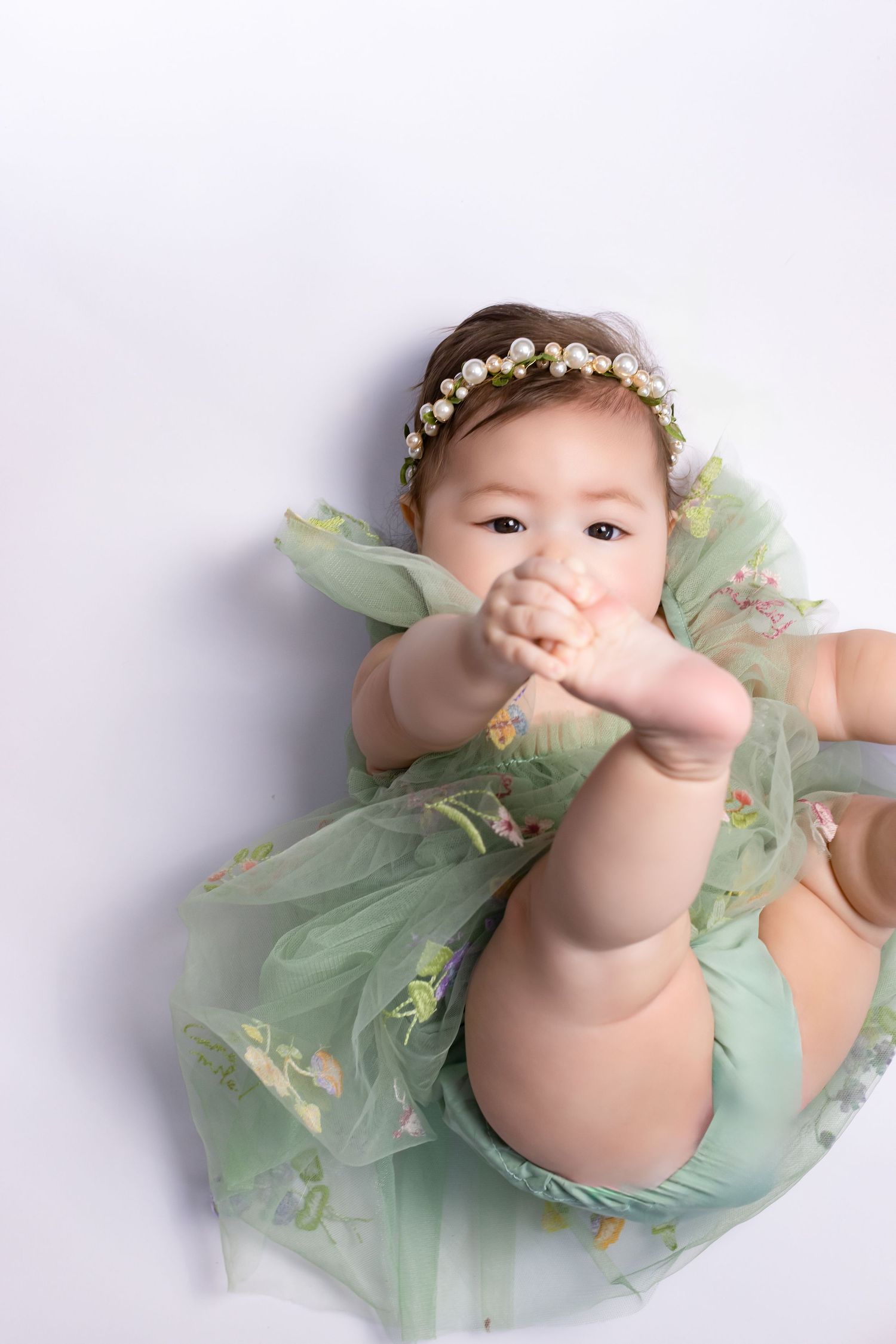 Adorable baby girl in green floral romper and pearl headband lying on white background chewing her hands.