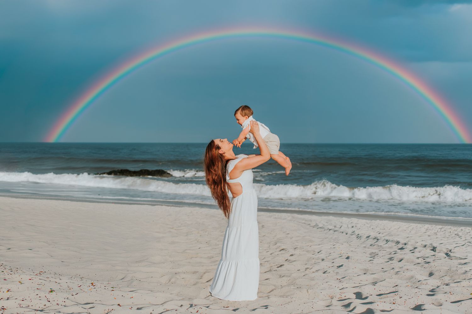 Mother in white dress lifting baby on beach under stunning full rainbow over ocean.