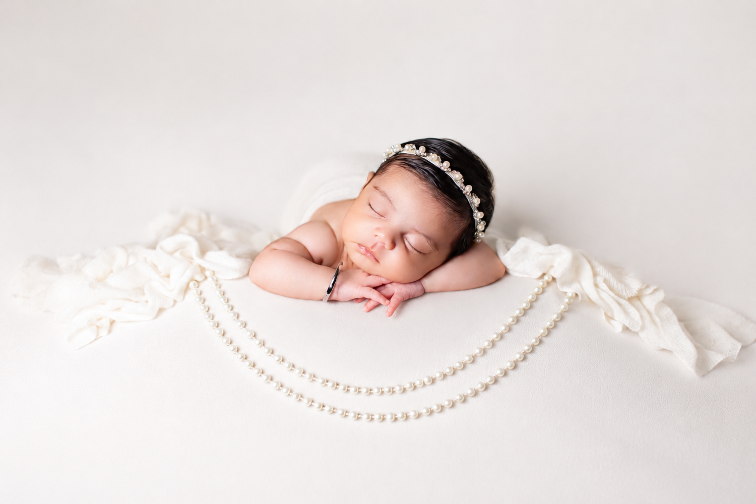 Sleeping newborn baby wearing a pearl headband, posed with hands under chin, surrounded by pearl necklaces on white backdrop.