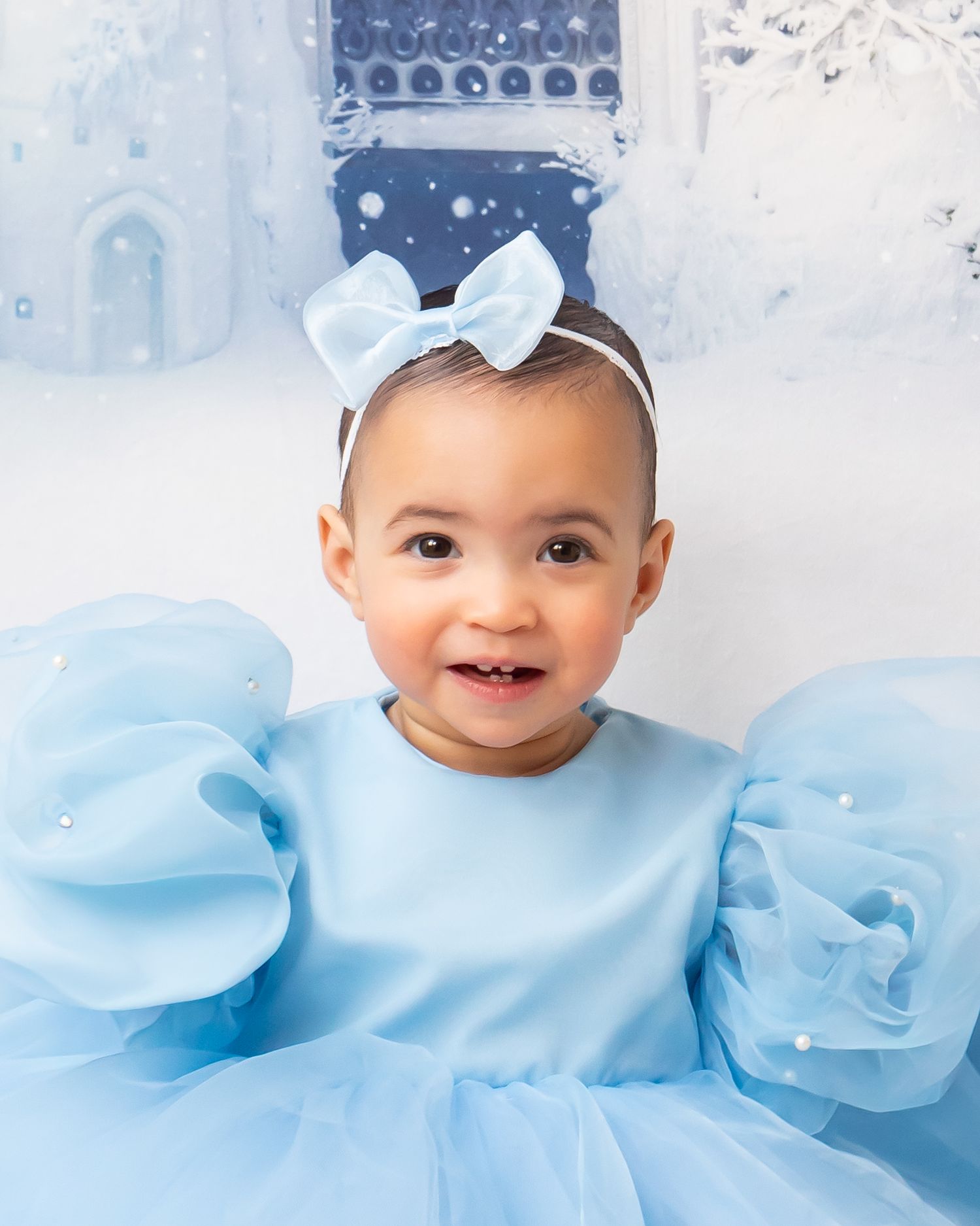 Happy baby girl in blue dress with bow headband smiles before a winter castle backdrop.