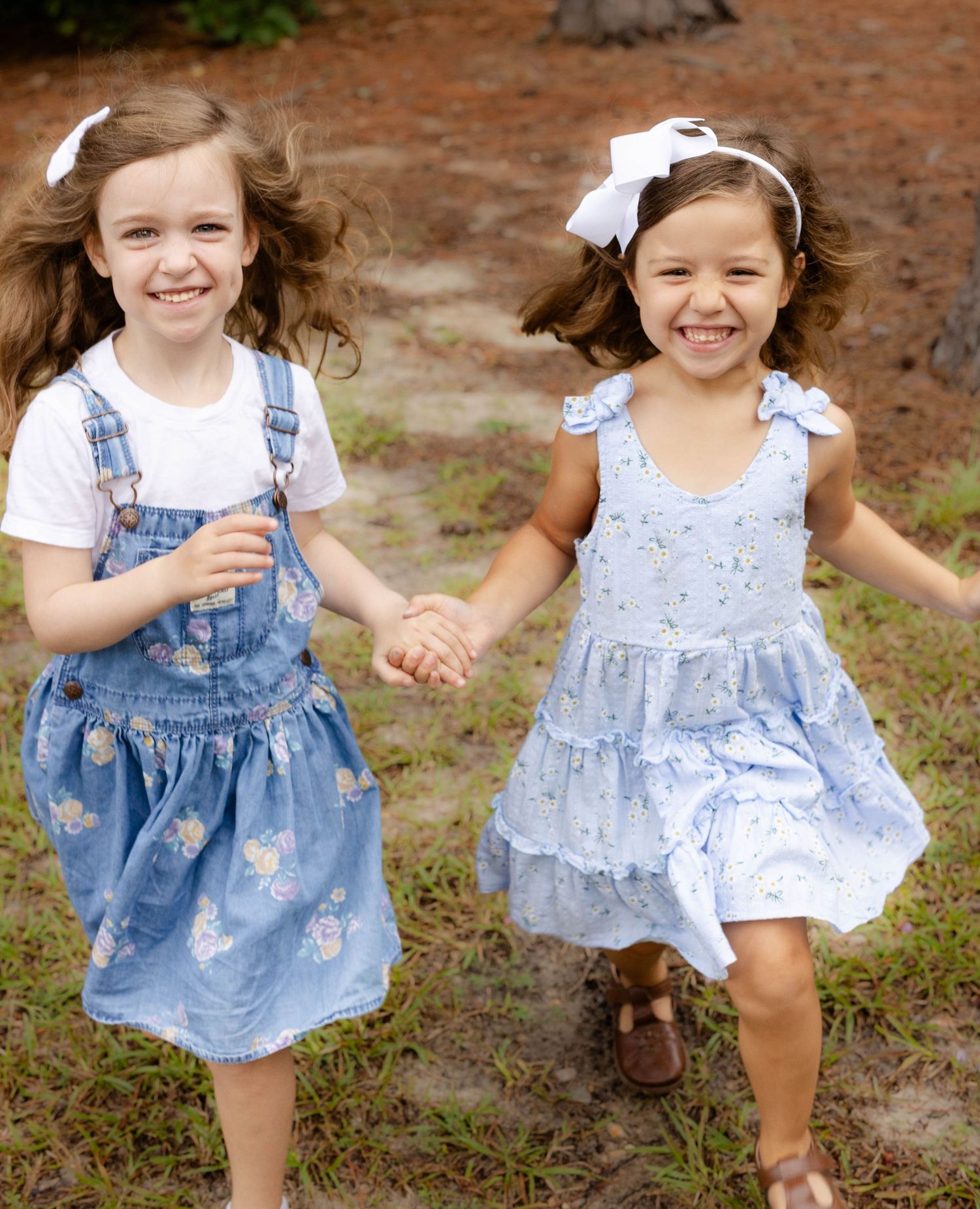 Two smiling girls in matching blue floral denim dresses and white hair bows hold hands while walking outdoors on a grassy path.