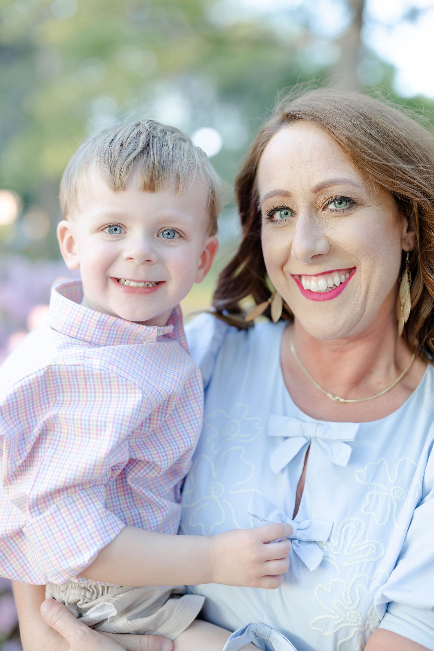 Smiling mother holds young blue-eyed boy in pink plaid shirt in outdoor portrait.