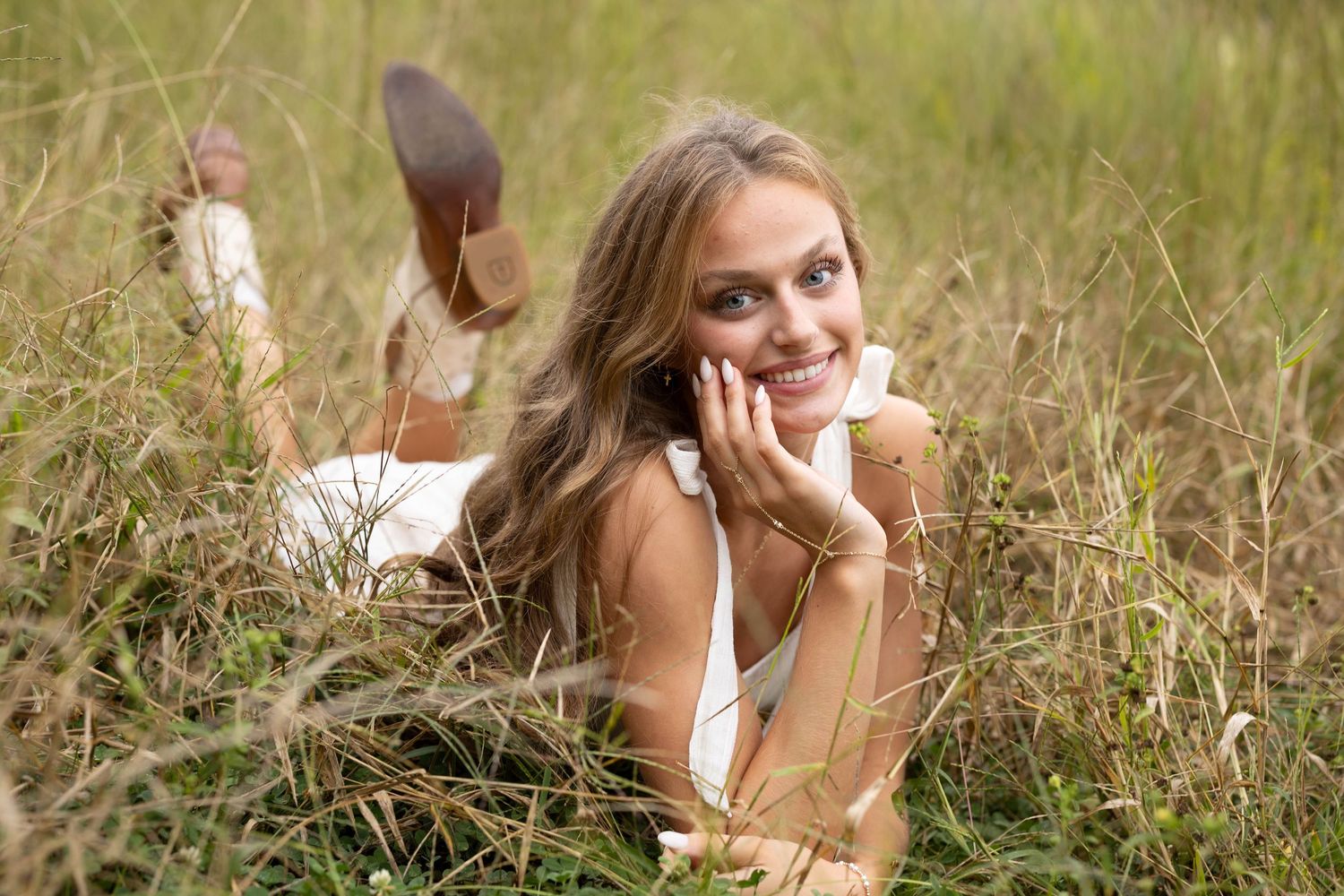 Smiling young woman with long hair lies on her stomach in tall green grass, resting her chin on her hand with feet kicked up.