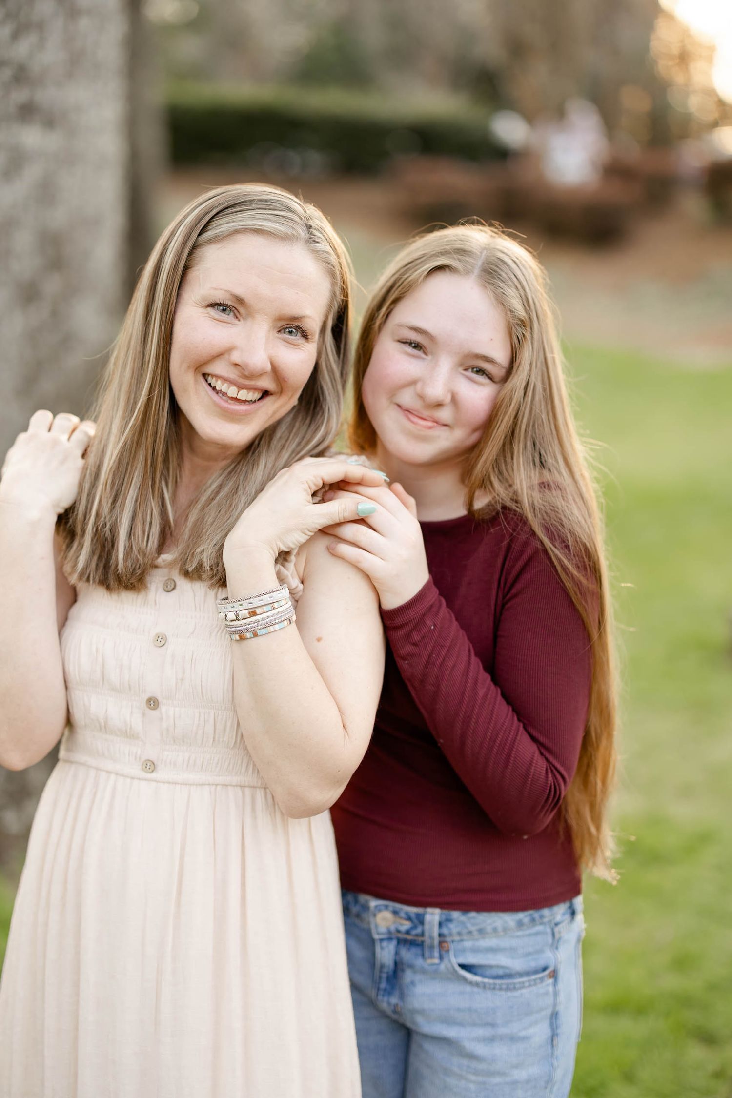 A smiling woman in cream dress and teenage girl in burgundy top pose together in a park.